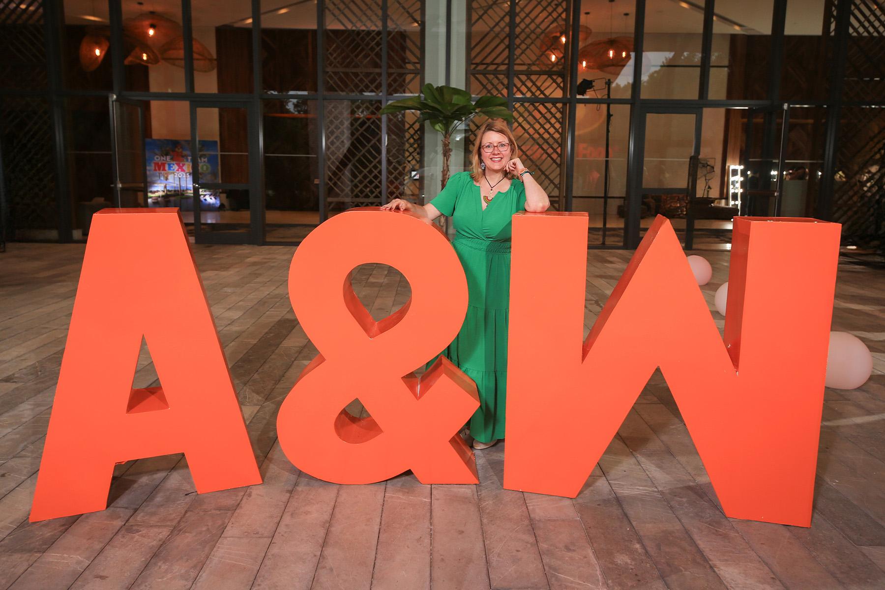 Betsy Schmandt wearing a green dress standing between large orange letters that spell "A&amp;W"